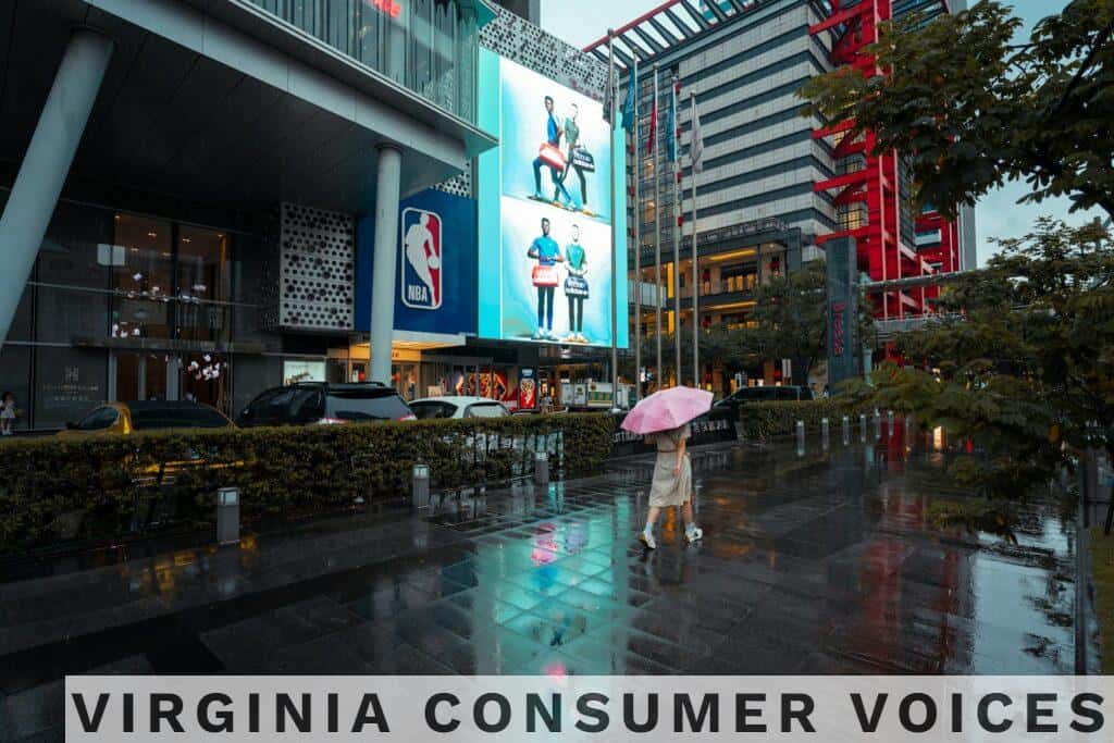 A woman walks with an umbrella past modern urban buildings and wet pavement reflecting city lights.